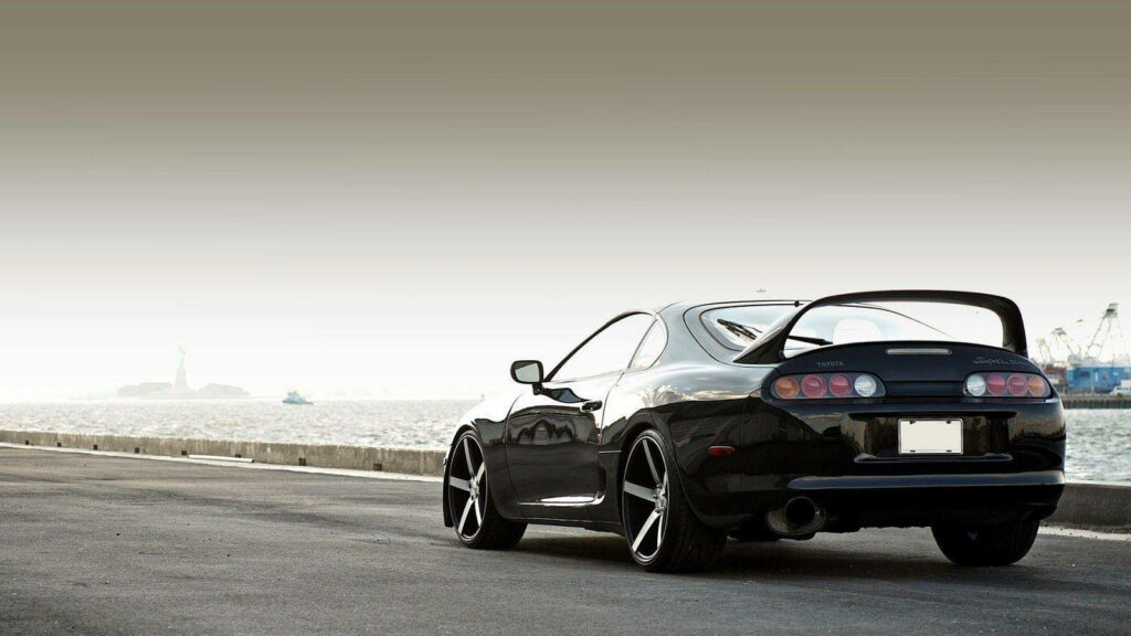 1994 Toyota Supra Mk4 black parked by waterfront with iconic rear wing and industrial harbor background Wallpaper