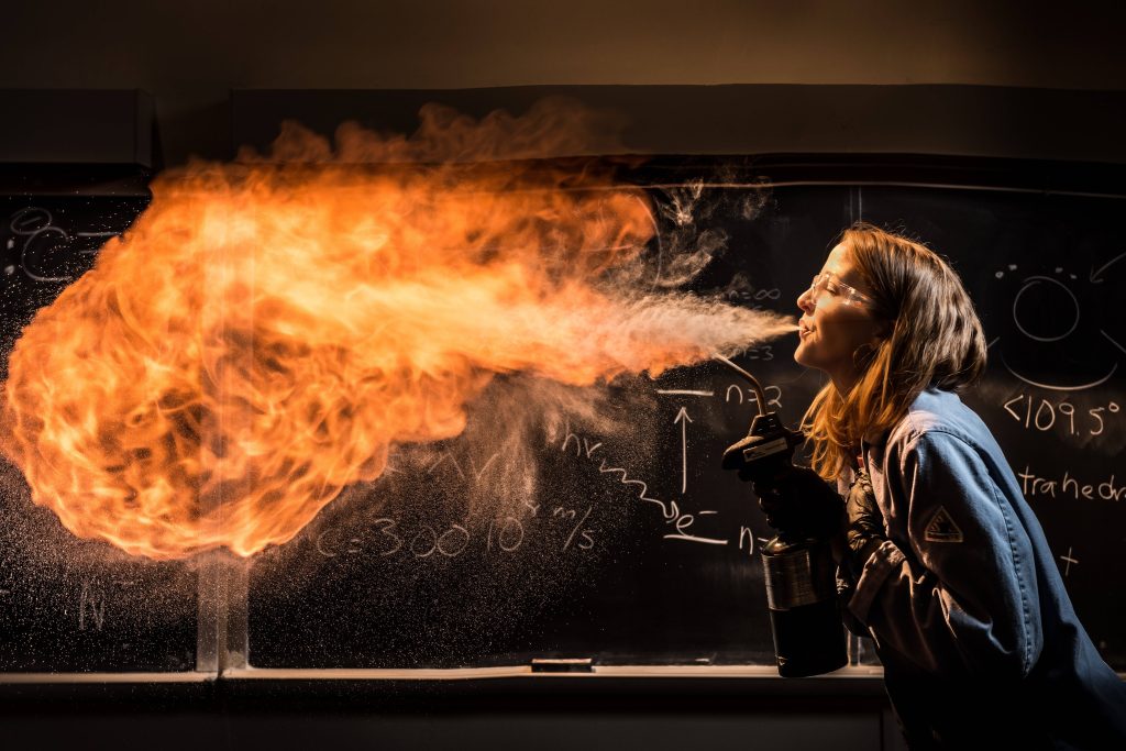Scientist woman in safety goggles creating a large fireball experiment with equations background Wallpaper