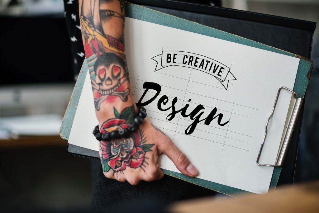 Tattooed arm with vibrant skull floral ink holding clipboard Be Creative Design wallpaper background