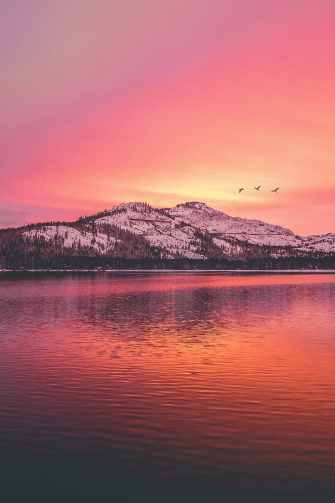 Snow-capped mountains and tranquil lake reflecting pink orange sunset with birds flying Wallpaper