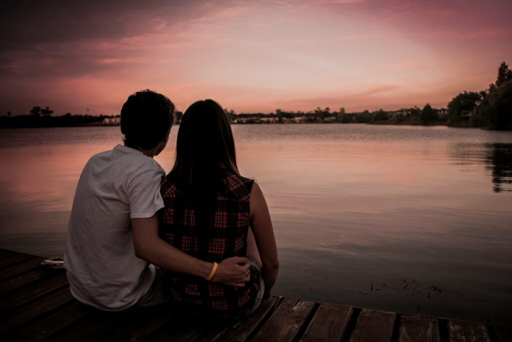 Couple sitting on wooden dock at lakeside sunset with peaceful calm water and warm pink sky Wallpaper