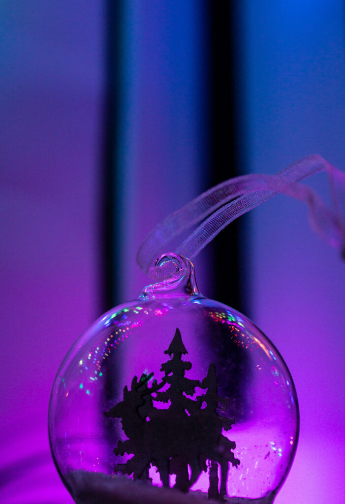 Glimpse into a Festive Winter Wonderland: Enchanting Glass Bauble with Reindeer and Pine Trees Gracefully Set in a Captivating Purple and Blue Gradient Glow Wallpaper
