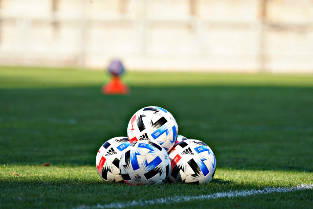 Stack of Adidas UEFA Champions League soccer balls on green pitch in bright natural daylight wallpaper