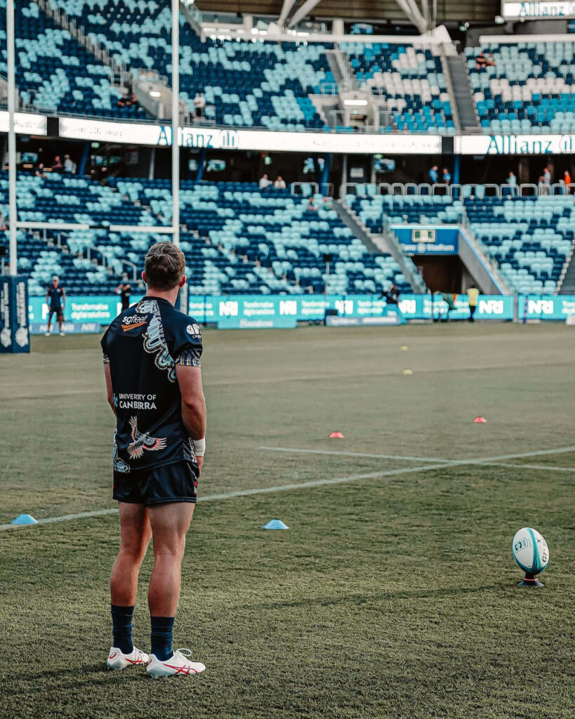 ACT Brumbies rugby player preparing to kick on Allianz Stadium field with blue seating background Wallpaper
