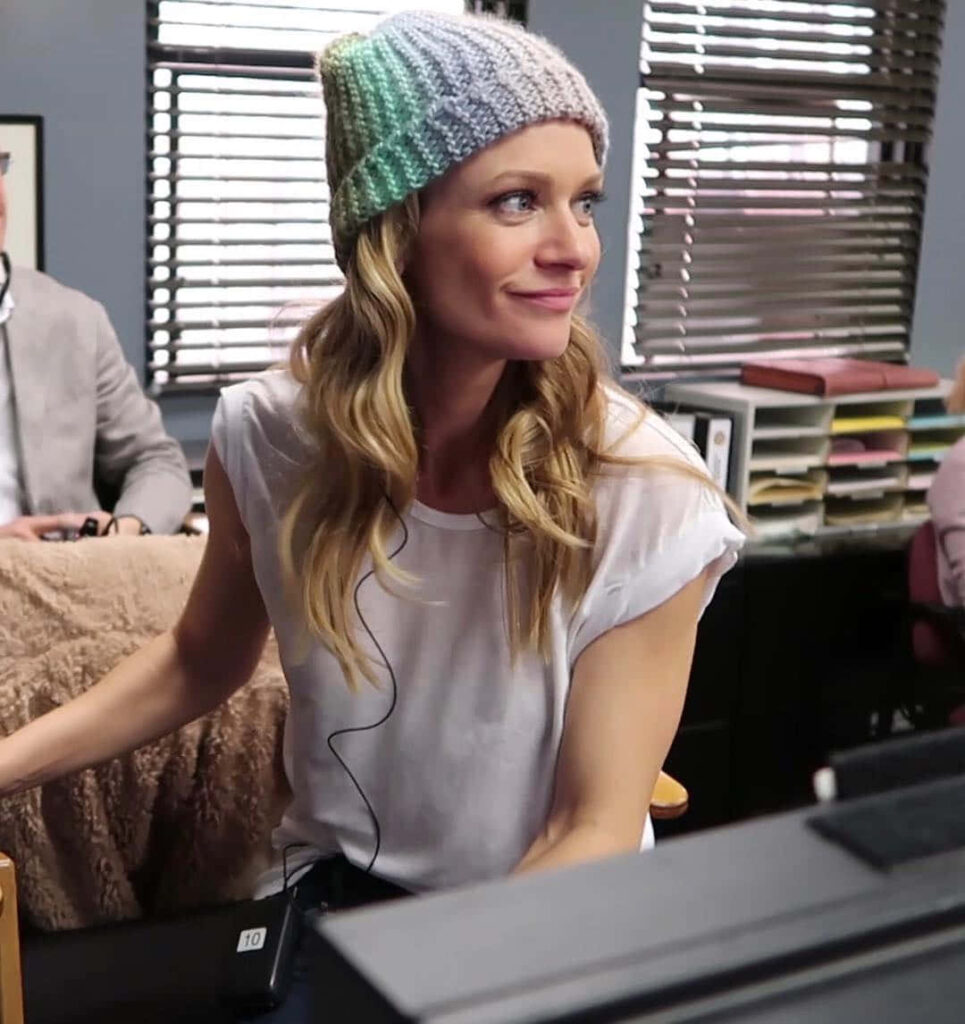 A.J. Cook in white t-shirt and colorful knit beanie seated in stylish casual workspace with wallpaper background