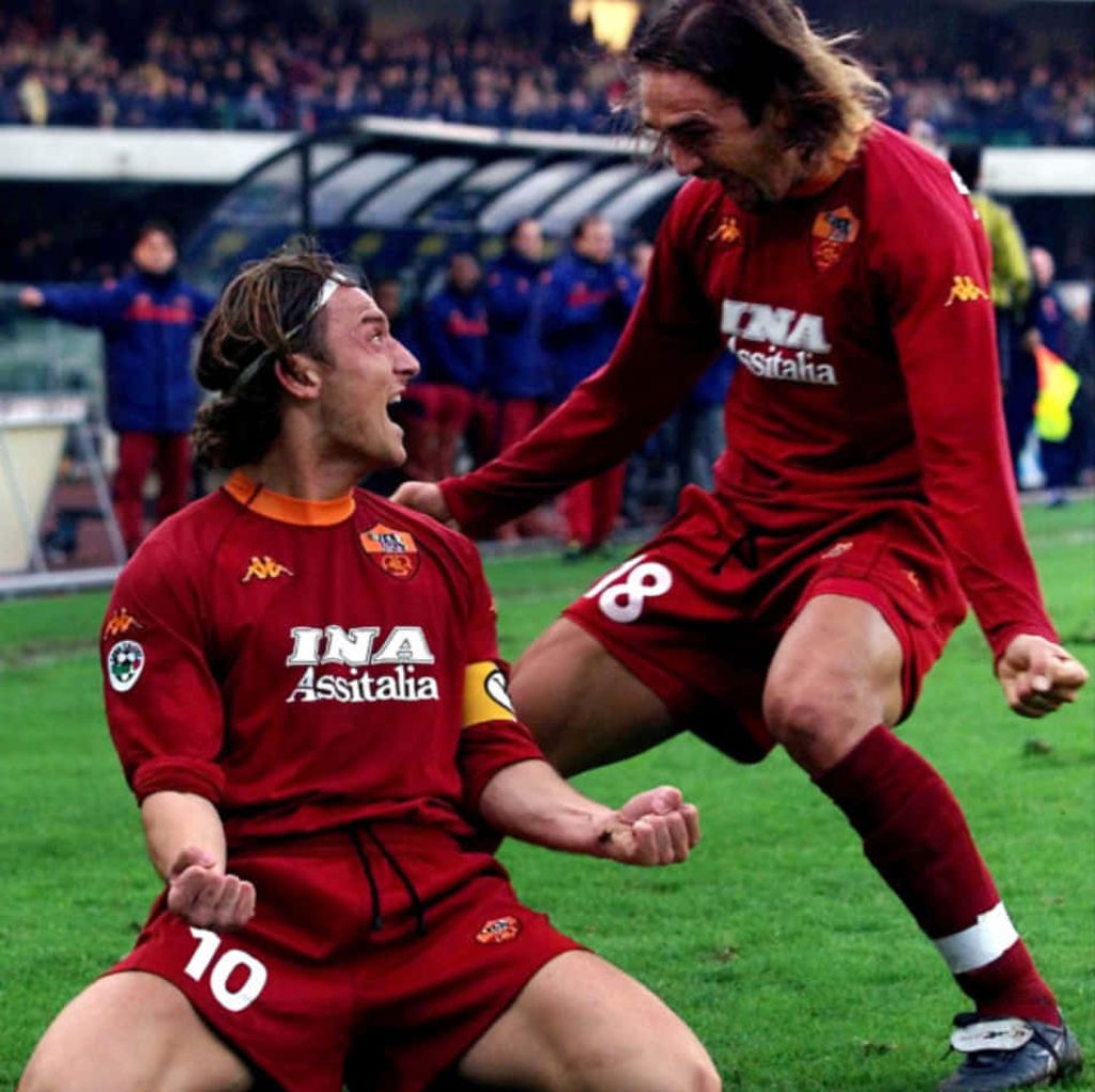 AS Roma legends Totti and Batistuta celebrate a goal in dynamic burgundy kits on green pitch Wallpaper