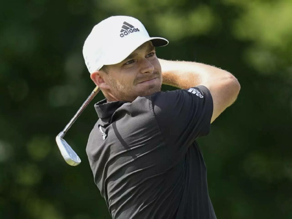 Aaron Wise focused golf swing wearing white Adidas cap black shirt outdoors lush green background Wallpaper