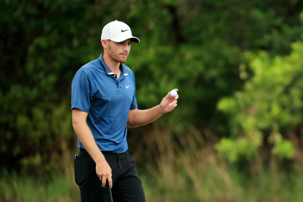 Aaron Wise confidently holds golf ball on course wearing blue polo white cap with green backdrop Wallpaper