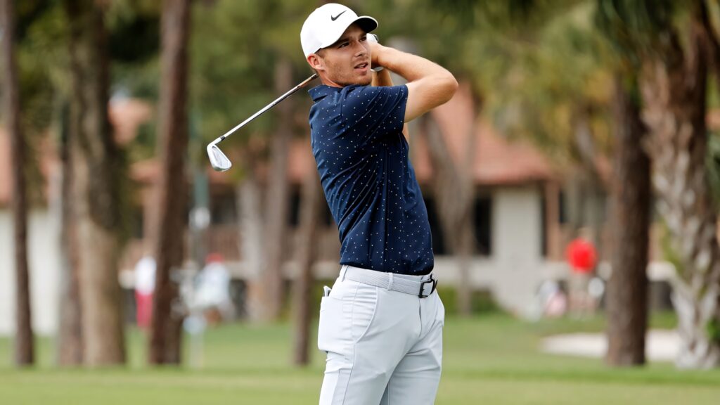 Aaron Wise mid-swing on lush golf course wearing navy polo and white pants focused golf wallpaper