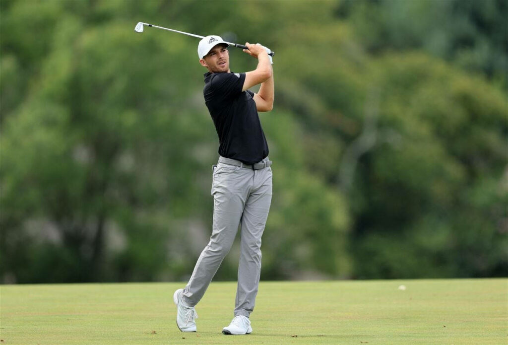 Golfer Aaron Wise mid swing on lush green course wearing professional golf attire against trees Wallpaper