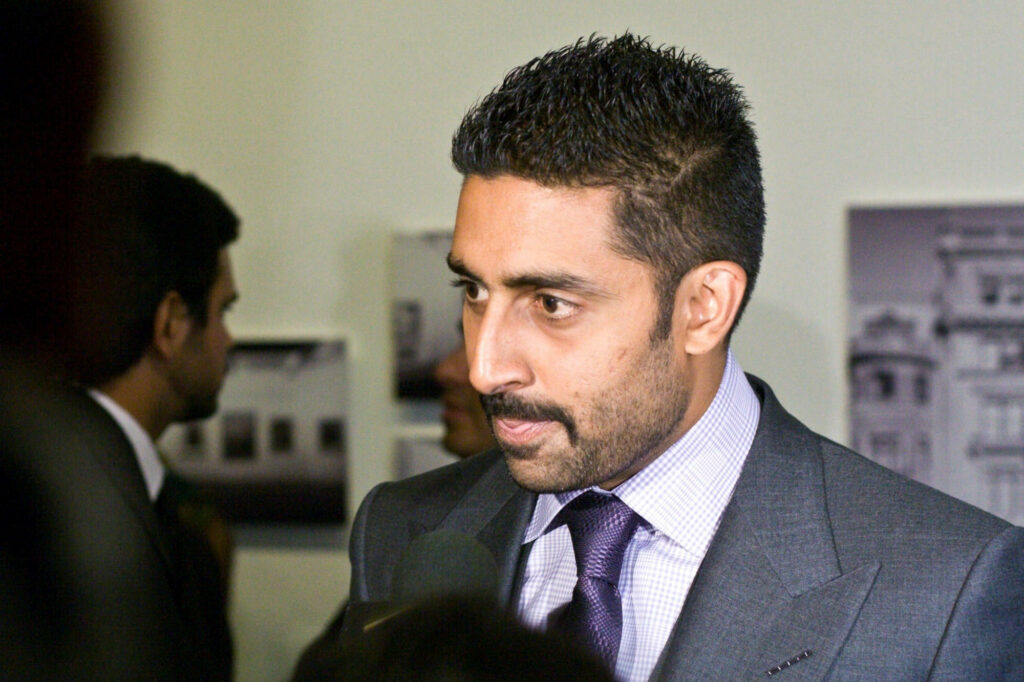 Abhishek Bachchan in sharp suit giving interview at formal event with elegant wallpaper backdrop