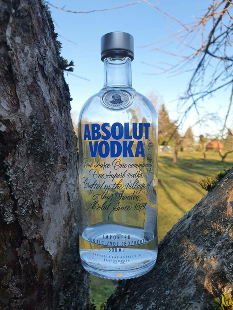 Absolut Vodka bottle framed by tree branches with sunlit park and blue sky background Wallpaper