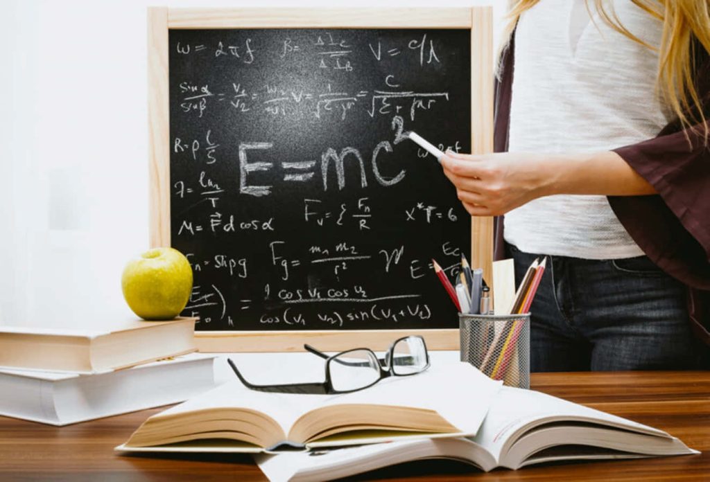 Teacher at blackboard with physics formulas open book apple glasses stationery on desk Wallpaper
