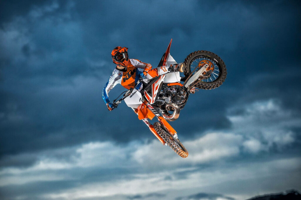 Dirt bike rider in orange white gear performing aerial stunt against dramatic cloudy sky wallpaper