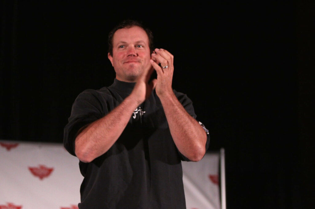 Adam Baldwin applauding enthusiastically on stage at event with decorative wallpaper background