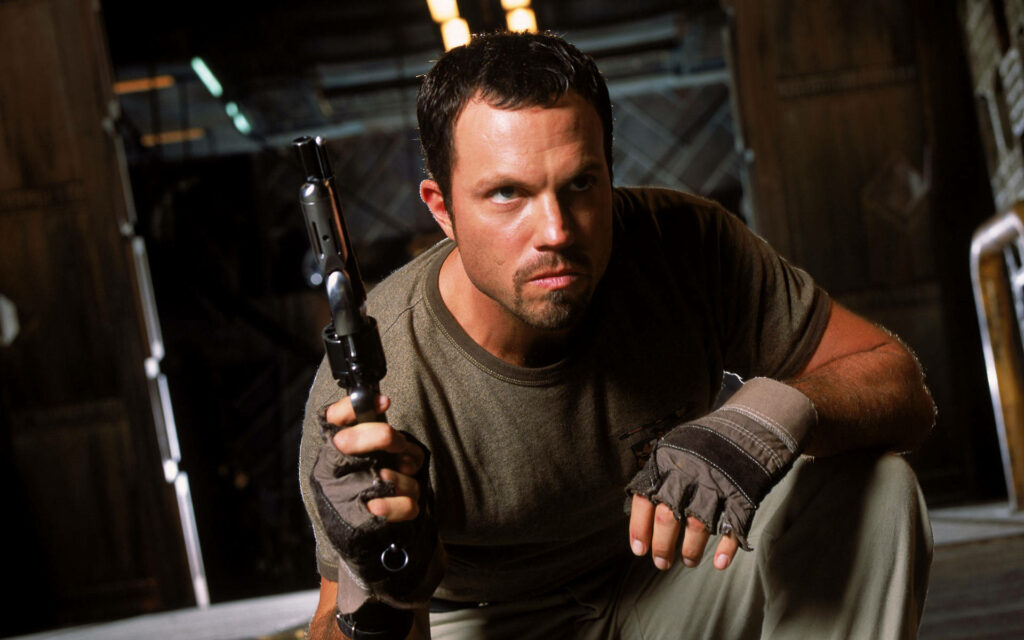 Adam Baldwin crouching with gun in dark industrial scene intense dramatic wallpaper background