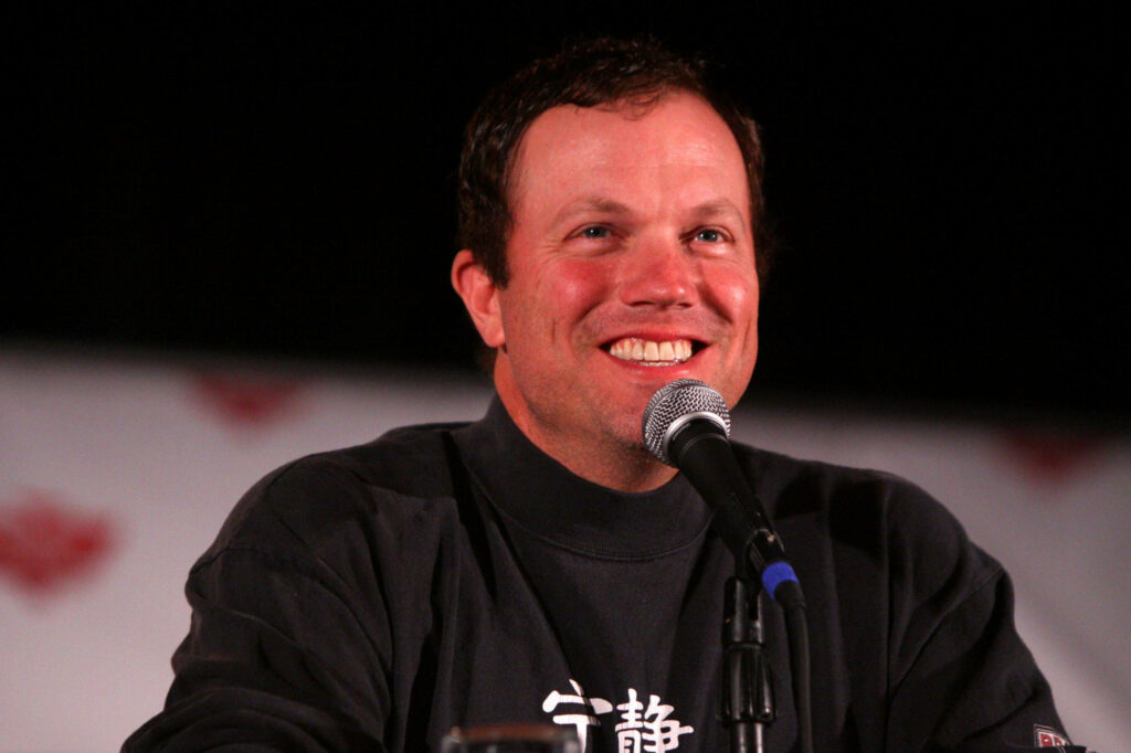 Adam Baldwin smiling and speaking on microphone at panel event lively engaging wallpaper background