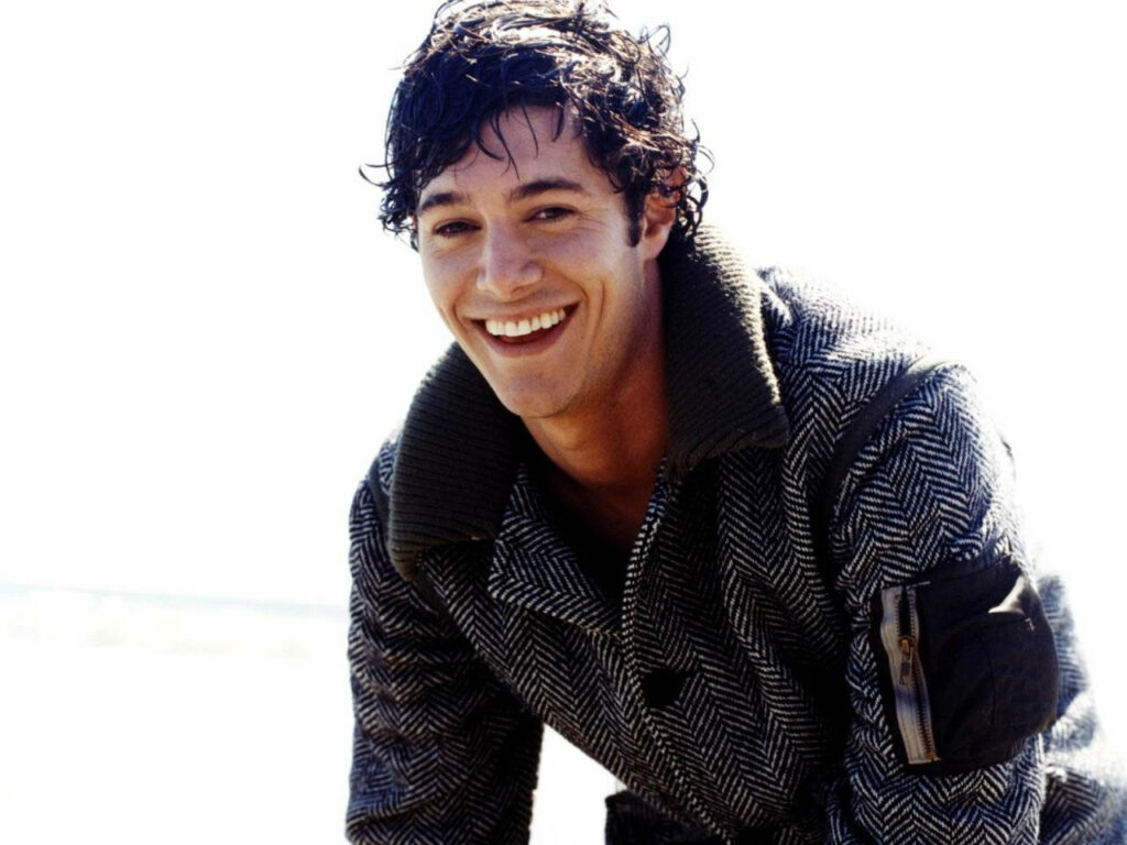 Adam Brody smiling with dimples wearing textured coat outdoors bright wallpaper background
