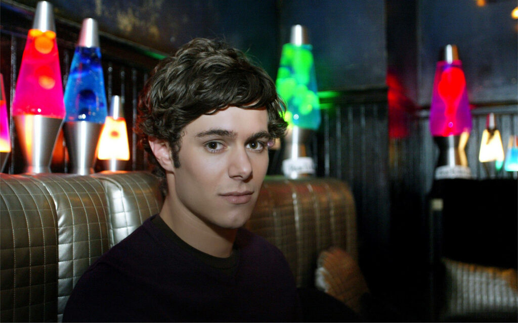 Adam Brody seated on bench in dim room with colorful lava lamps retro relaxed wallpaper background