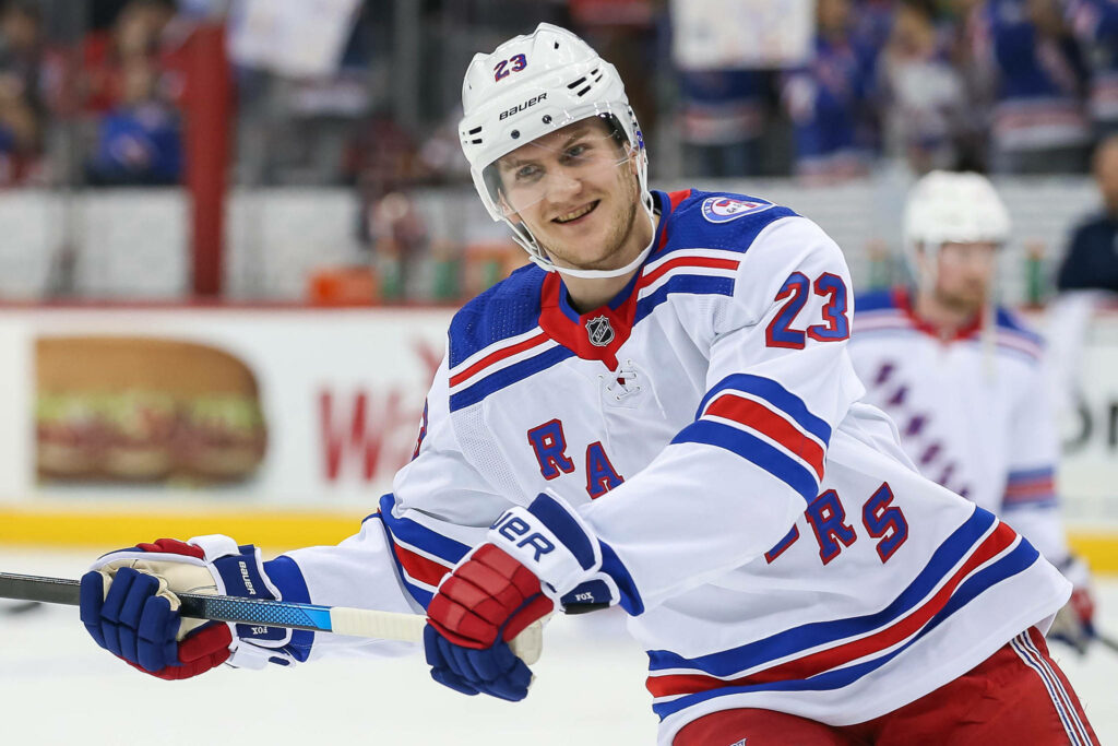 Adam Fox smiling in white New York Rangers uniform showing confidence on ice hockey rink Wallpaper