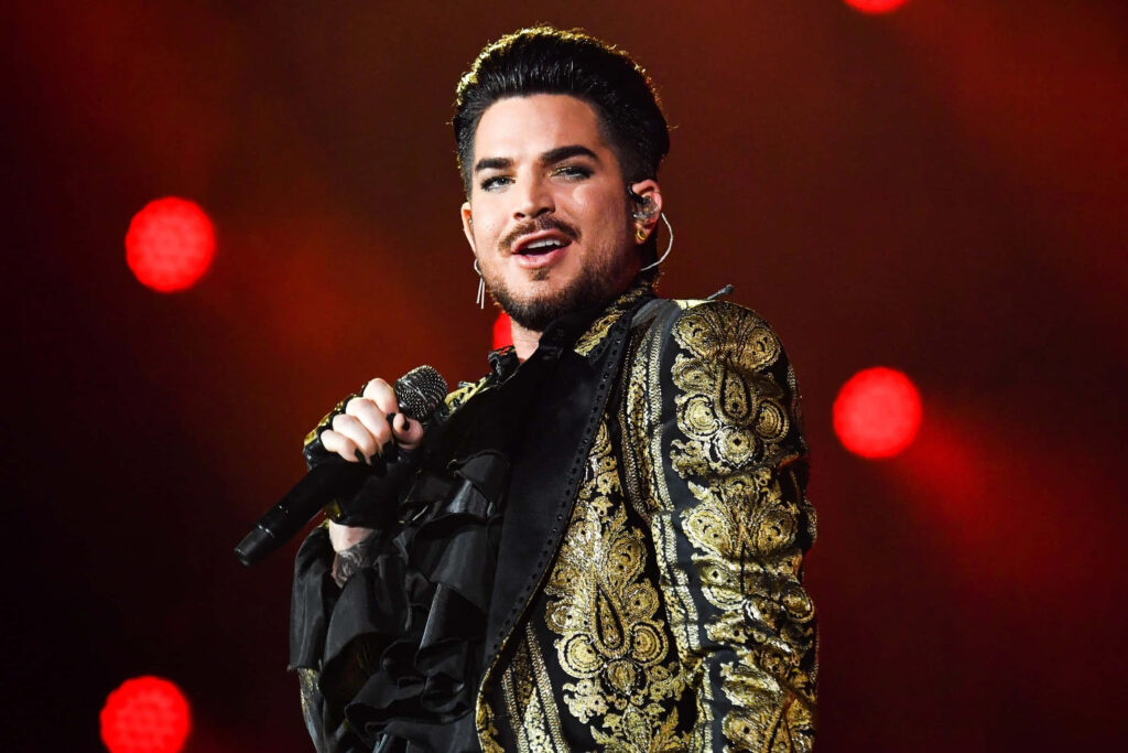 Adam Lambert on stage wearing black gold embroidered jacket singing with red lights background Wallpaper