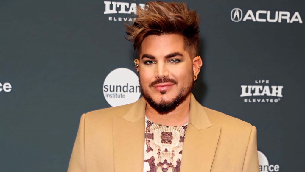 Adam Lambert stylish in tan blazer and patterned shirt at event with sponsor logo backdrop Wallpaper