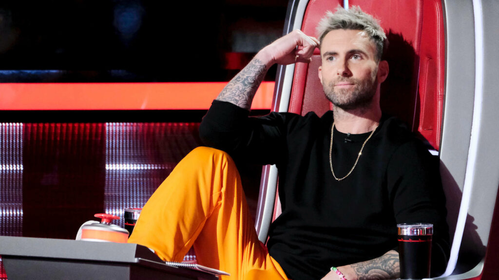 Adam Levine in black and orange attire, deep in thought in The Voice's iconic red chair. Wallpaper