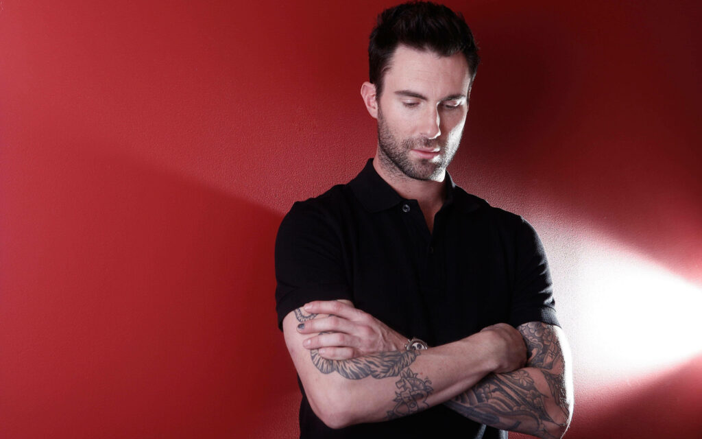 Adam Levine in a classic black outfit against a vibrant red backdrop, showcasing his tattoos. Wallpaper