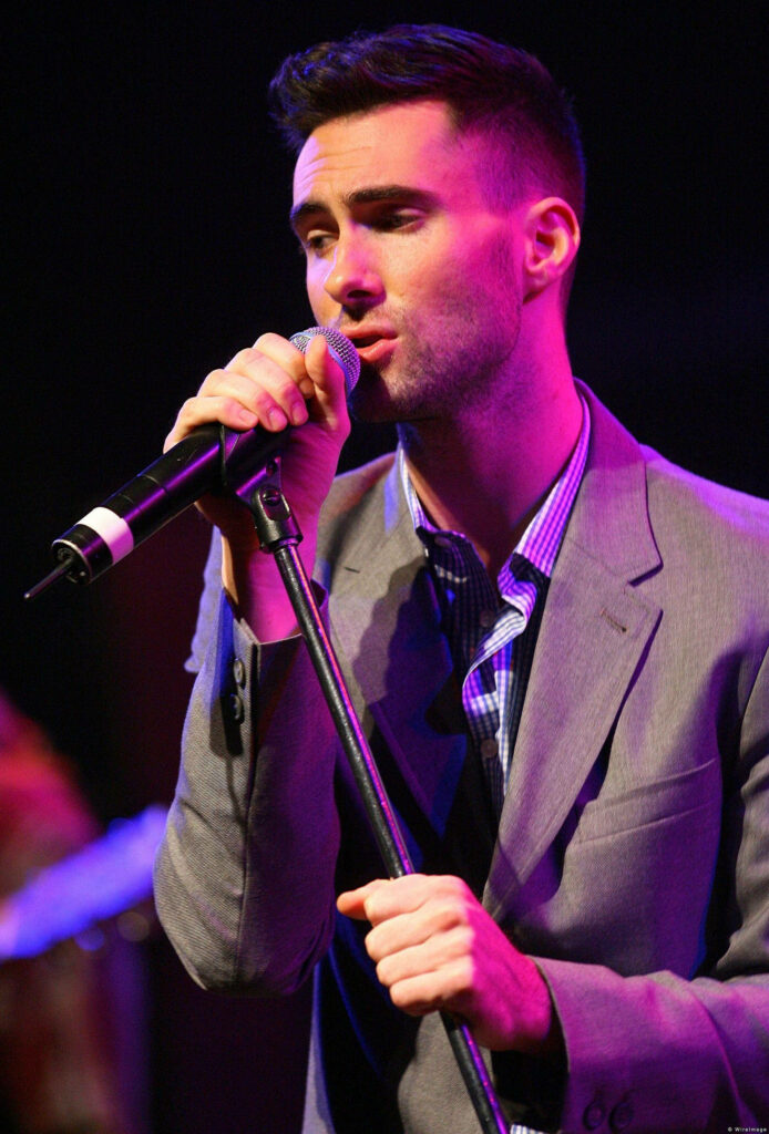 Adam Levine performing passionately in a stylish grey suit under vibrant stage lights wallpaper.