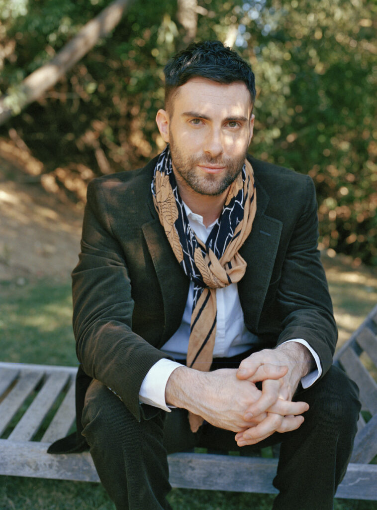 Adam Levine relaxing outdoors in stylish outfit with patterned scarf against serene nature backdrop. Wallpaper