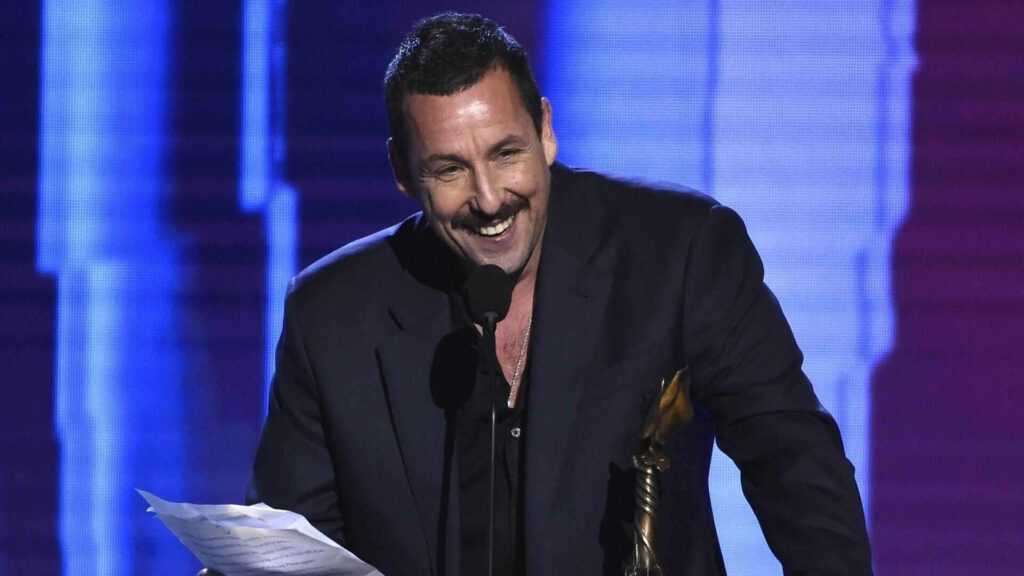 Adam Sandler smiling and speaking at microphone holding paper on stage with vibrant blue lighting Wallpaper