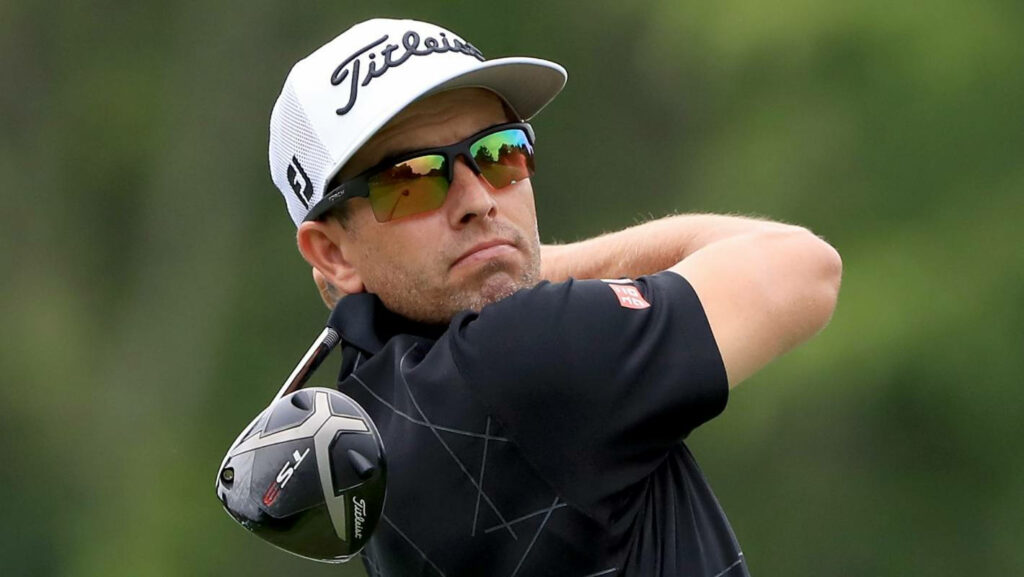 Adam Scott mid golf swing wearing multicolored sunglasses golf cap lush green course background Wallpaper