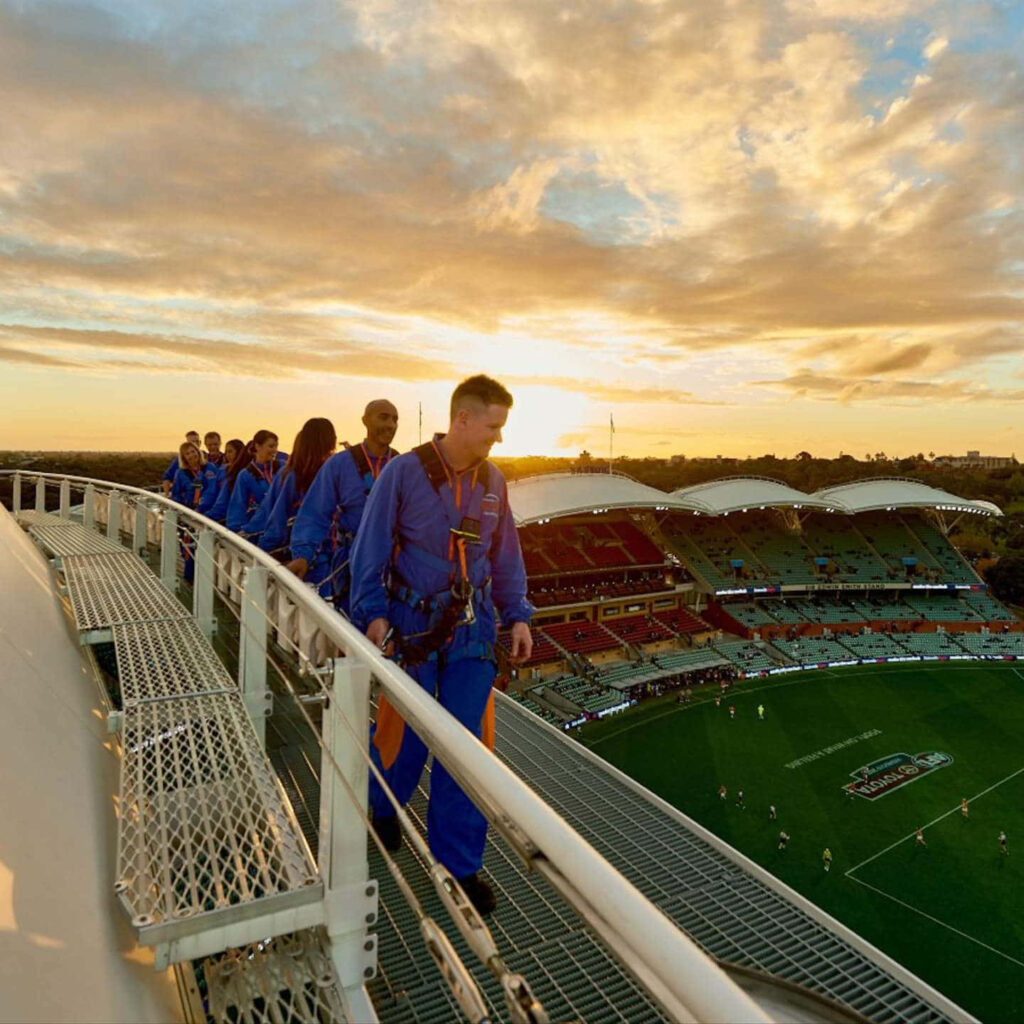 Adventurers in blue suits walk rooftop Adelaide Oval sunset golden light stadium clouds wallpaper