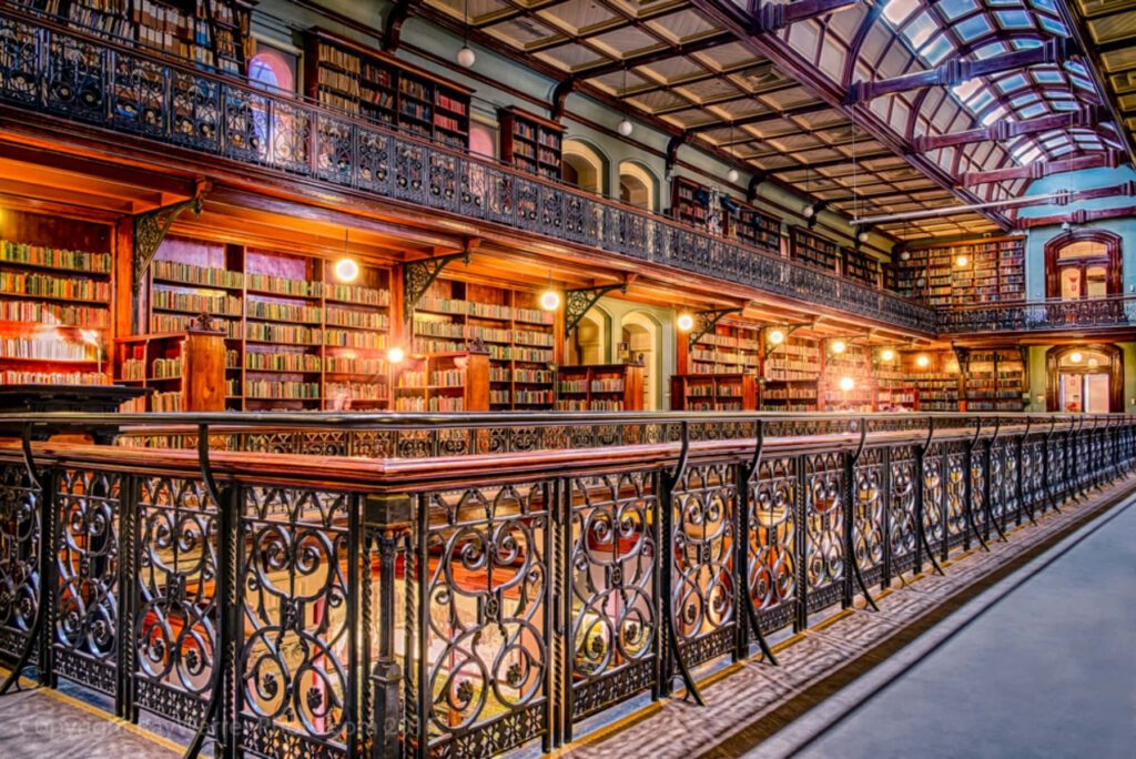 Adelaide State Library interior with ornate iron railings colorful bookshelves and warm lighting Wallpaper