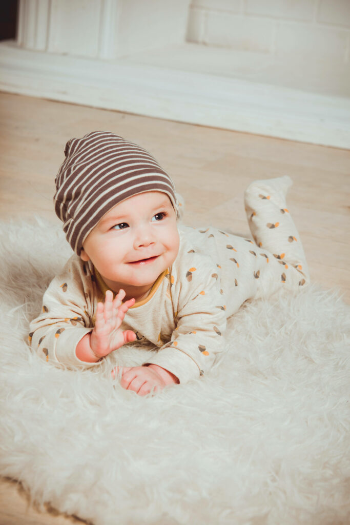Smiling baby boy in striped beanie and cozy pajamas lying on soft white rug background Wallpaper