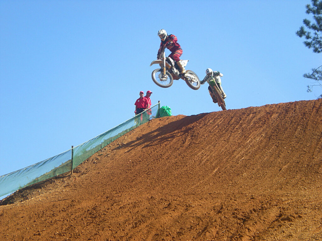 Two motocross riders mid-air over sunlit dirt track with clear blue sky action wallpaper