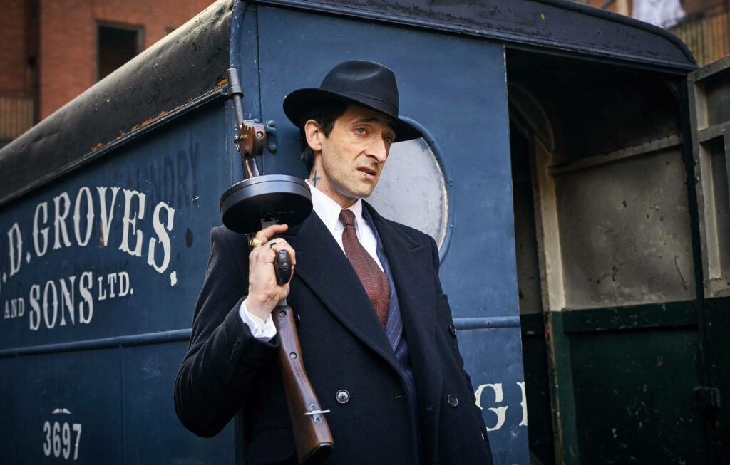 Adrien Brody as Peaky Blinders character in classic suit hat holding vintage firearm by vintage car Wallpaper