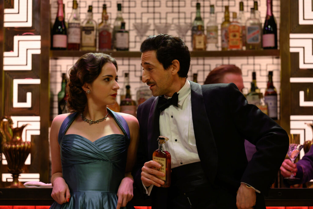 Adrien Brody in tuxedo leaning at bar with woman in blue dress vintage bottles background Wallpaper