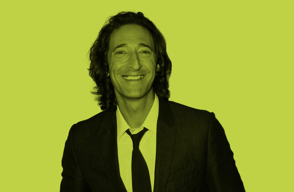 Adrien Brody smiling in classic suit on bold yellow wallpaper background showcasing charisma