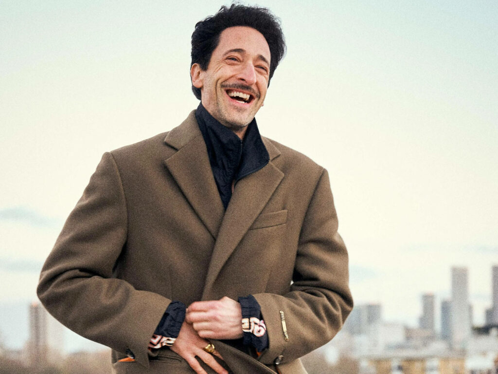 Adrien Brody smiling outdoors wearing brown coat with cityscape background wallpaper