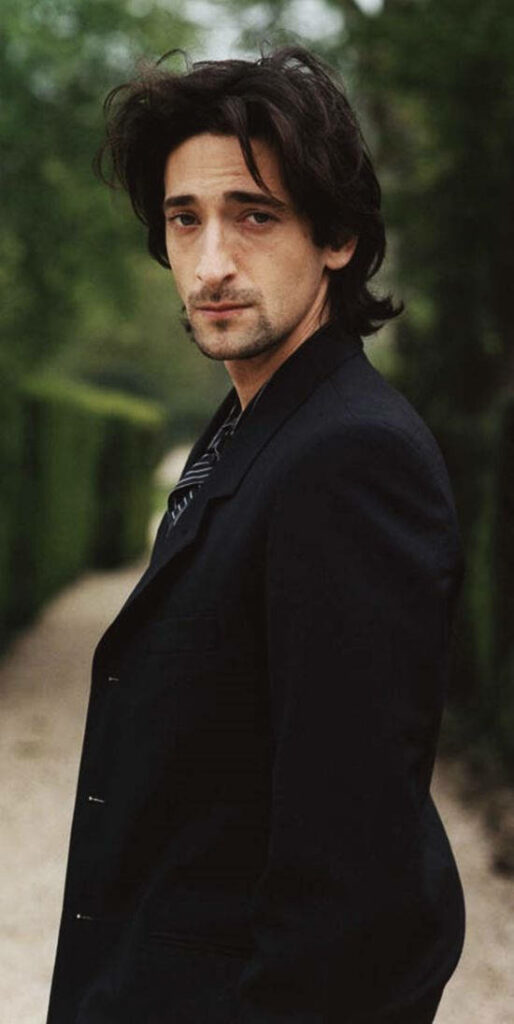 Adrien Brody in dark suit on lush green outdoor walkway stylish contemplative portrait background Wallpaper
