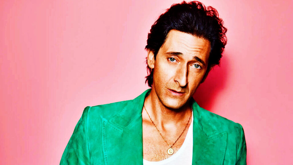 Adrien Brody in green jacket and white shirt posing against vibrant pink stylish wallpaper background