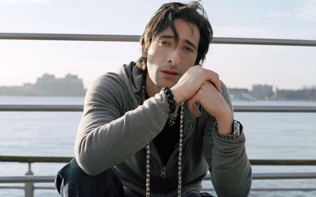 Adrien Brody sitting thoughtfully by the peaceful waterfront in a calm outdoor wallpaper background
