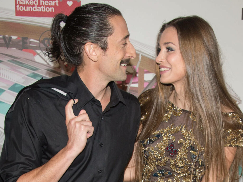 Adrien Brody and Lara Lieto smiling elegantly on red carpet with event logo backdrop wallpaper