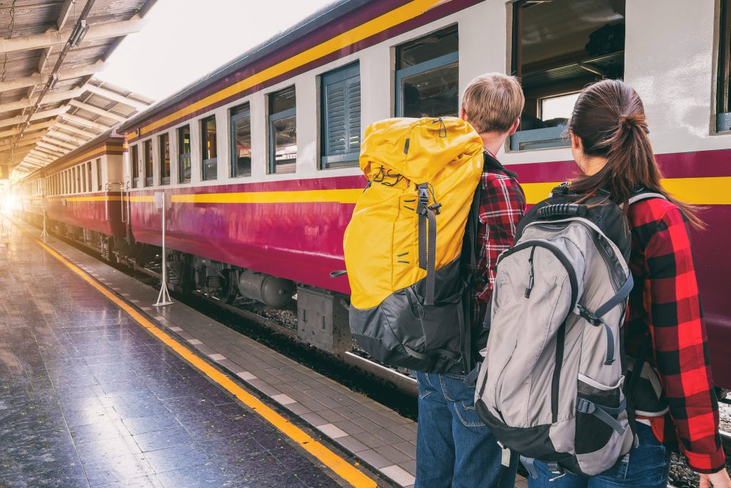 Two backpackers in red plaid shirts await a maroon cream train on a sunny station platform Wallpaper