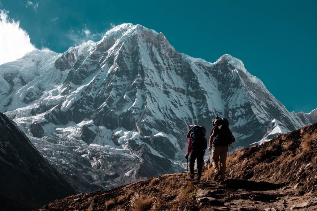 Two tourists trekking a rugged mountain path with snow-covered peak under clear blue sky background Wallpaper