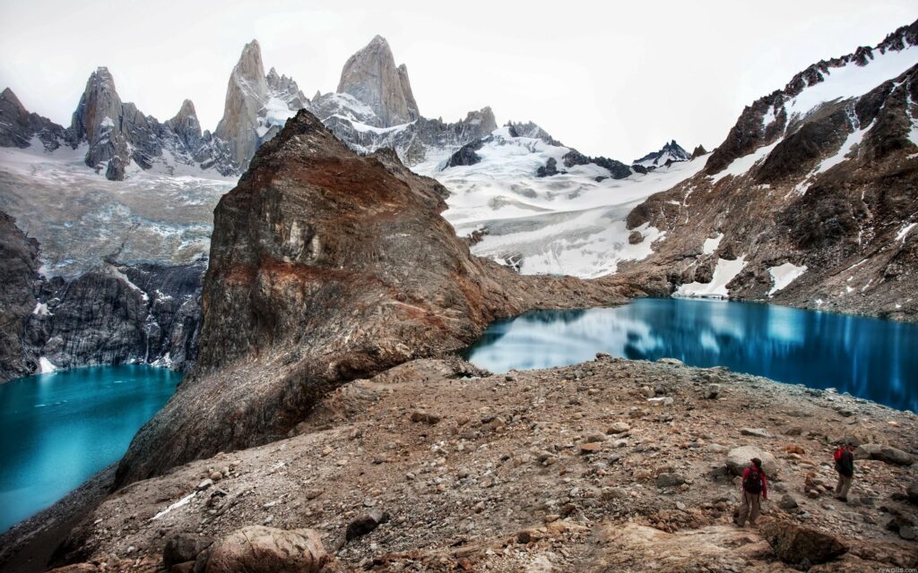 Rugged climbers trekking rocky terrain by blue lakes with snow-capped mountains background Wallpaper