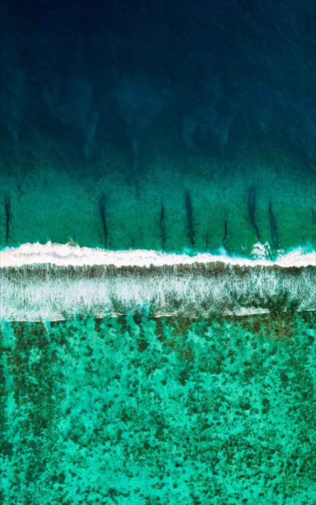 Aerial view of ocean with deep blue water turquoise shallows and foamy white wave on shore Wallpaper