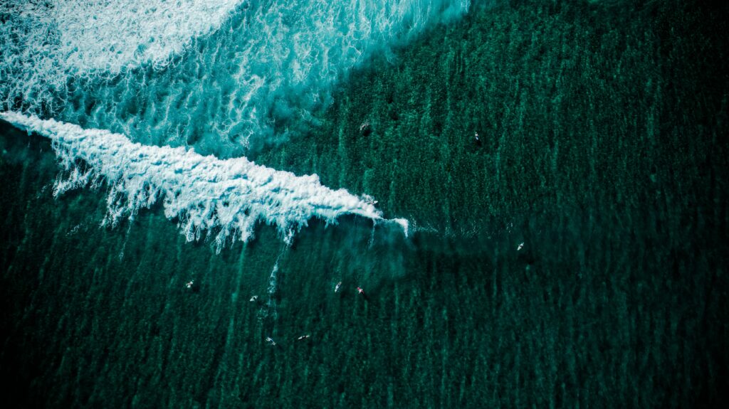 Aerial view of turquoise wave breaking over deep green ocean with surfers and boats background Wallpaper