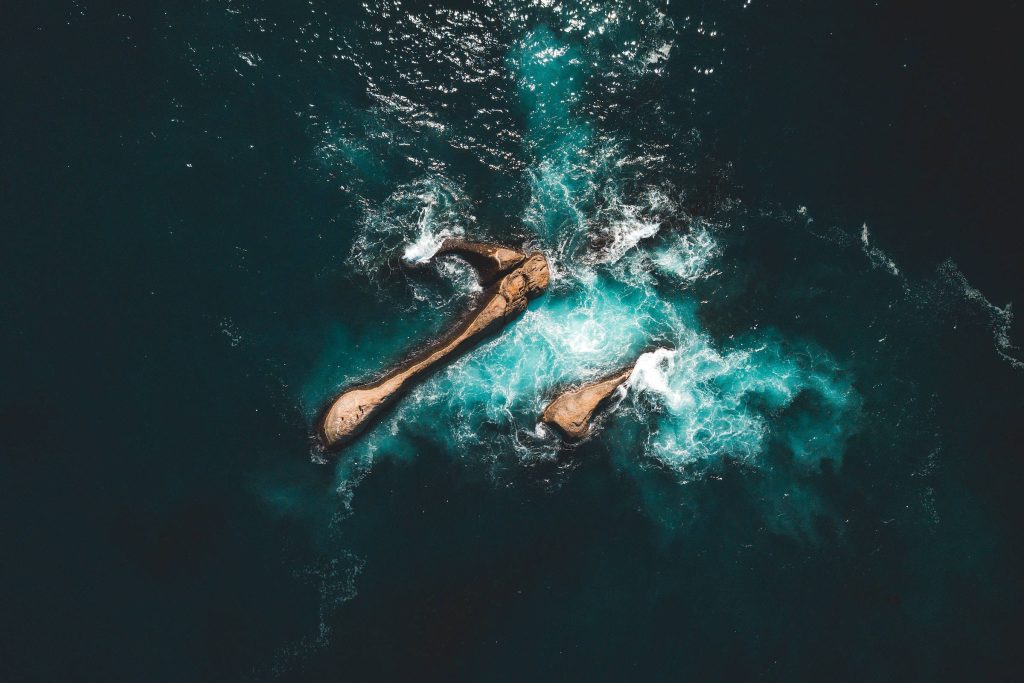 Aerial view of rugged rocks in deep blue ocean with vibrant white waves crashing beautifully Wallpaper